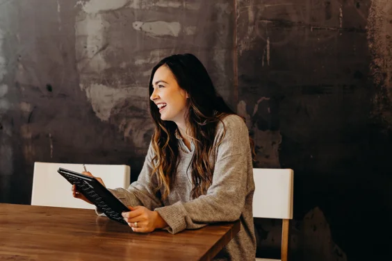 Woman with brown long hair sat at a desk, holding a notepad and laughing.